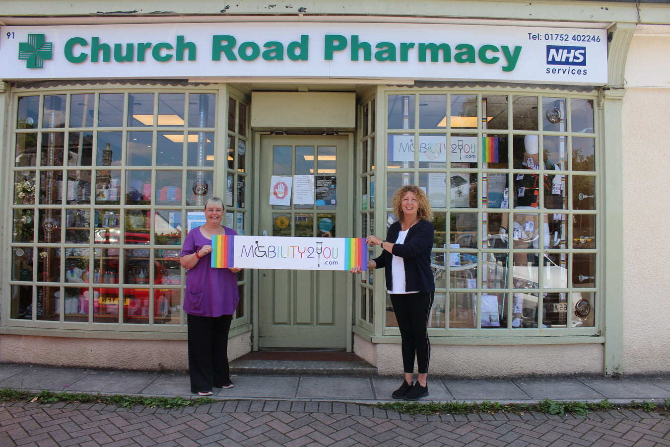 2 staff members holding a Mobility2You sign outside Church Road Pharmacy.